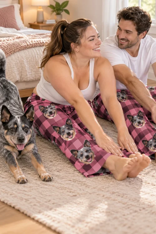 Couple stretching on a bedroom rug in pink plaid cattle dog pajama pants while their heeler does a play bow