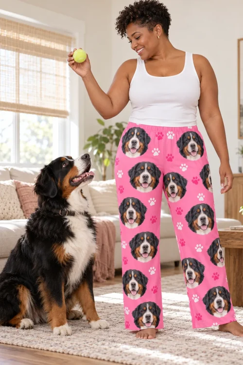 Woman holding a tennis ball above a seated Bernese, wearing pink paws pajama pants printed with Bernese faces