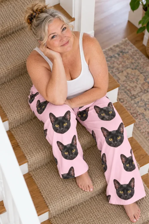 Woman with blonde hair resting her chin on her hand on carpeted stairs, in blush pink black cat pajama pants