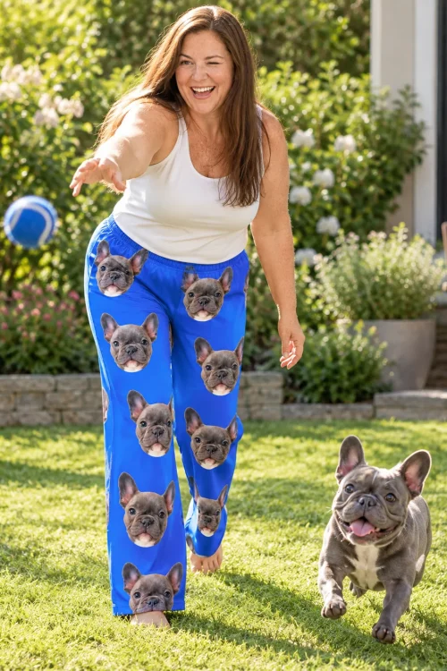 Woman tossing a ball to a running French bulldog in the yard, wearing blue pajama pants with its face