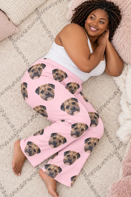 Woman lying on a cream rug, seen from above, in blush pink pajama pants printed with border terrier faces