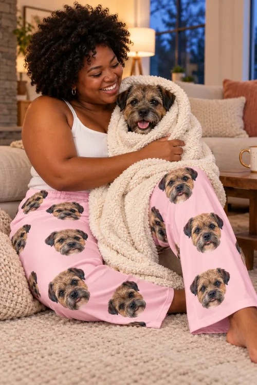 Smiling woman on a shag rug wrapping her border terrier in a cream blanket by the fireplace, blush pink pajamas