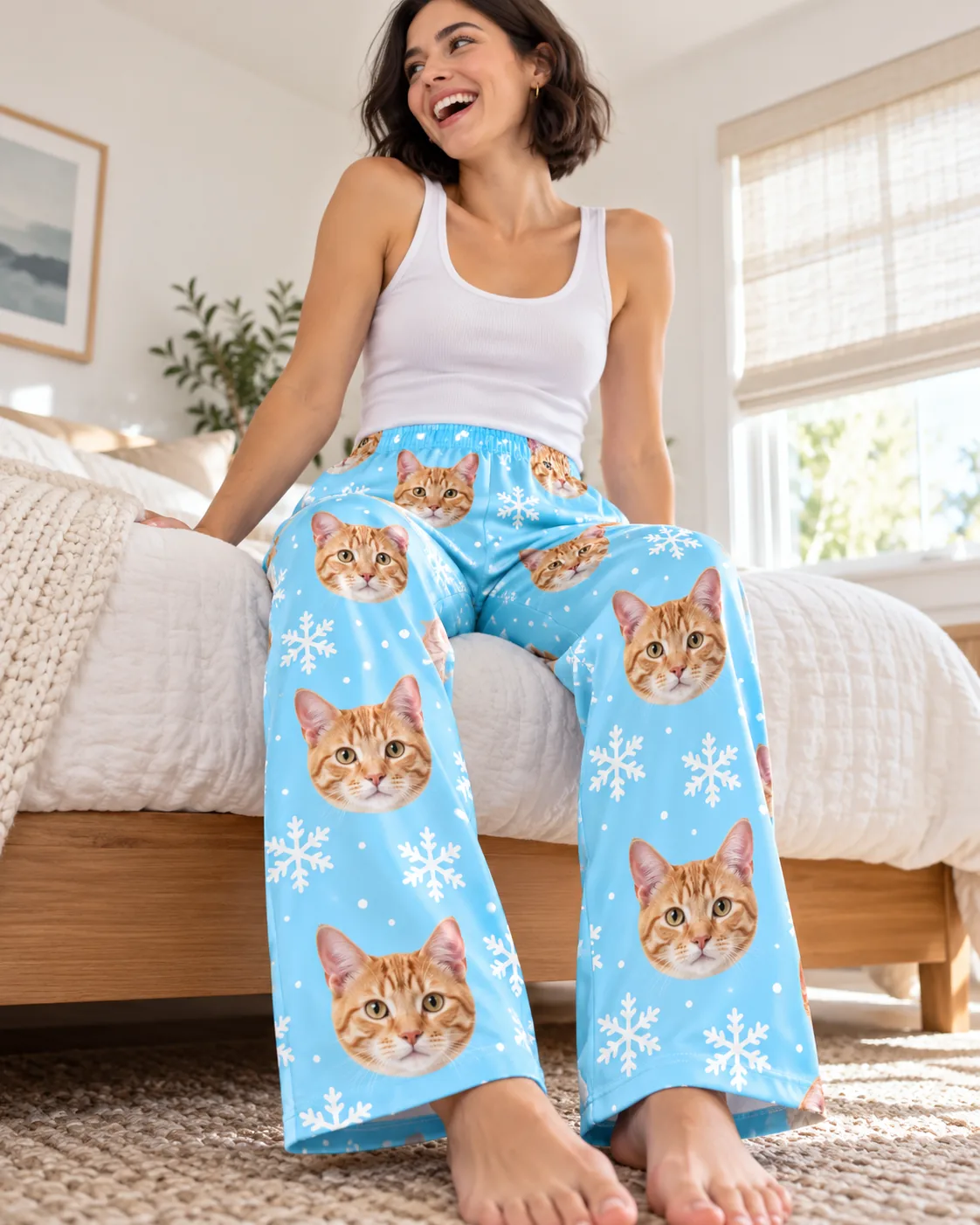 Women's Custom Cat Face Pajama Pants — snowflake detail | GenzOutfit Laughing woman sitting on the edge of her bed in snowflake cat pajama pants with tabby faces