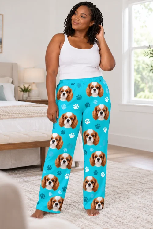 Woman in a white tank top tucking her hair back in a bright bedroom, in teal paws pajama pants with cavalier faces