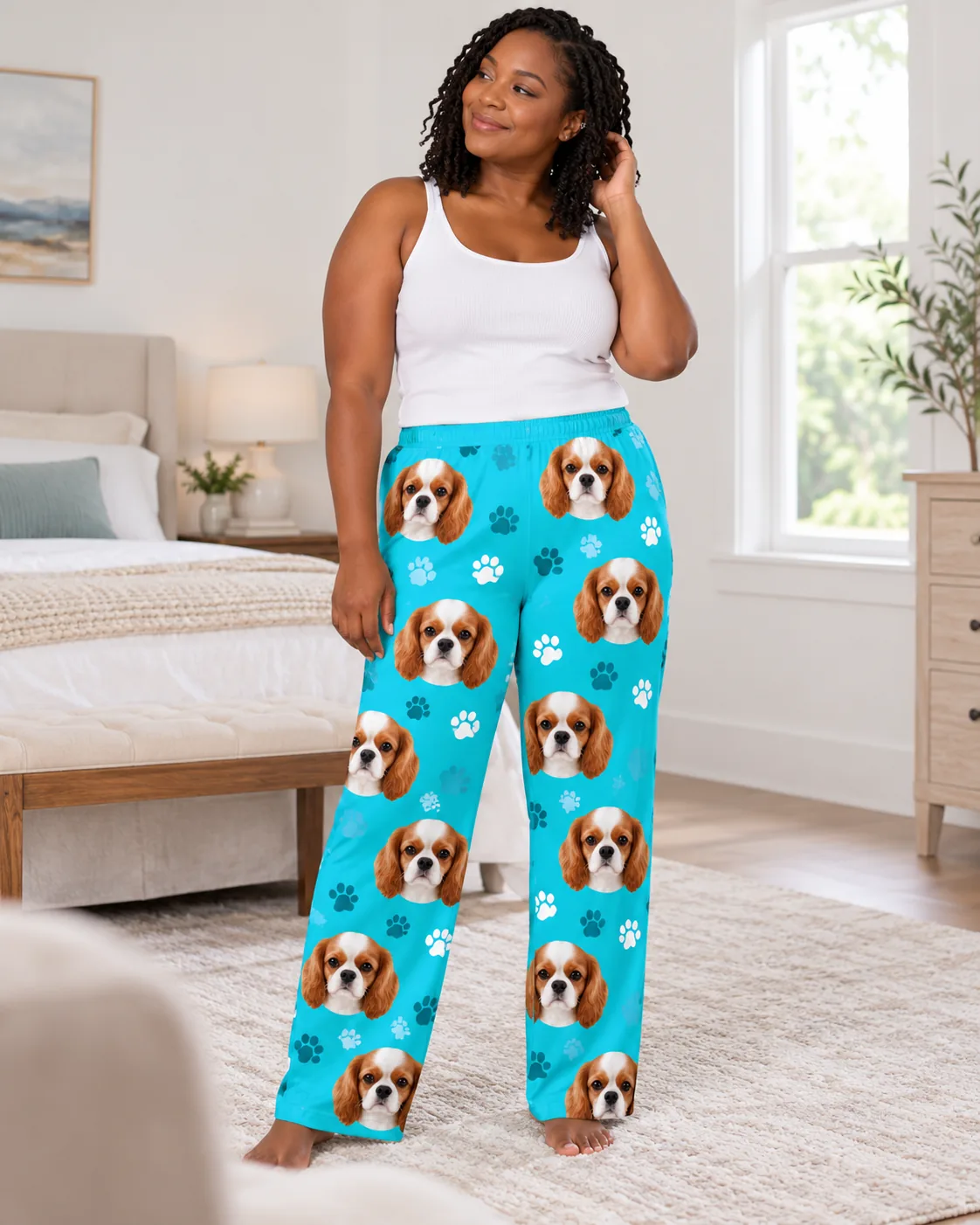 Woman in a white tank top tucking her hair back in a bright bedroom, in teal paws pajama pants with cavalier faces