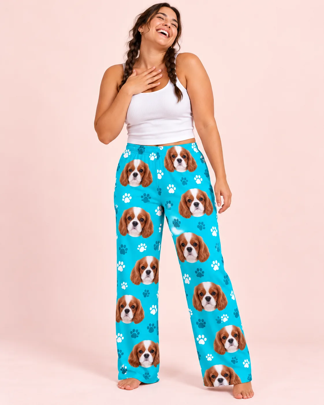 Laughing woman with two braids and a hand on her chest, in teal paws cavalier pajama pants against a blush pink wall