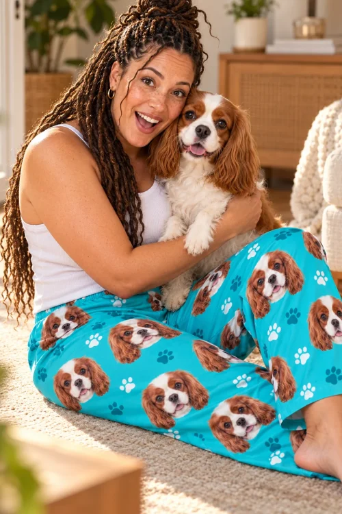 Woman with long twists hugging her Blenheim cavalier on a sunny rug, in teal paws pajama pants