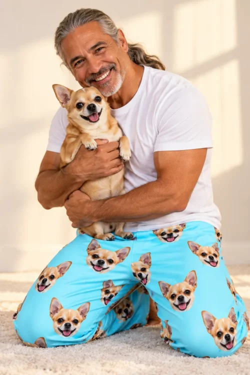 Man kneeling on a cream rug hugging a chihuahua, in light blue pajama pants printed with its face