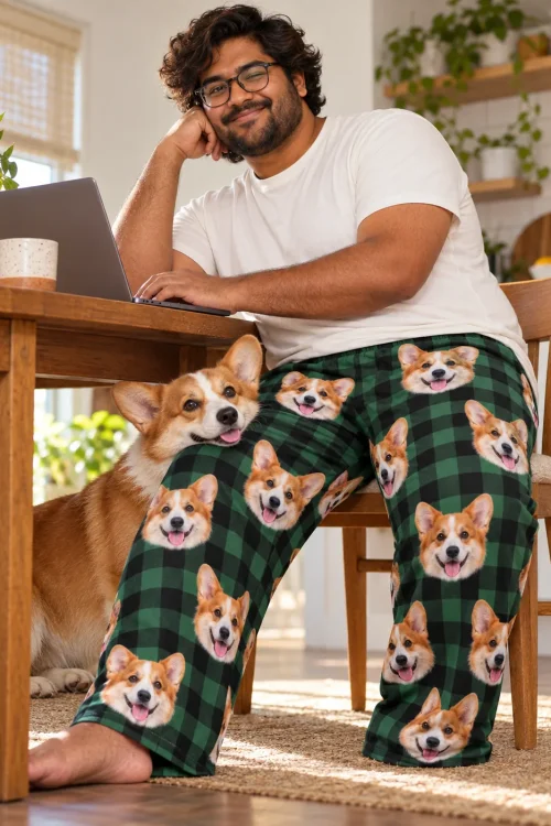 Man in glasses at a laptop in green plaid corgi pajama pants while his corgi rests its chin on his knee