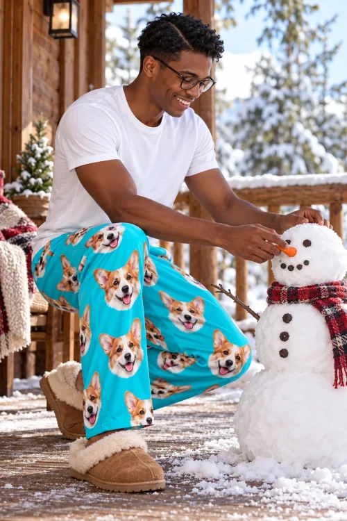 Man in glasses building a snowman on a snowy cabin porch in teal corgi pajama pants and sheepskin slippers