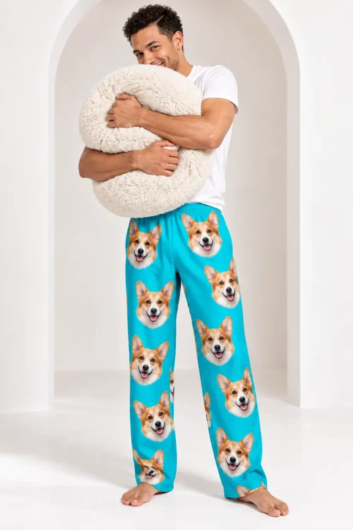 Smiling man hugging a fluffy cream pillow in teal corgi pajama pants covered in corgi faces, white studio arch