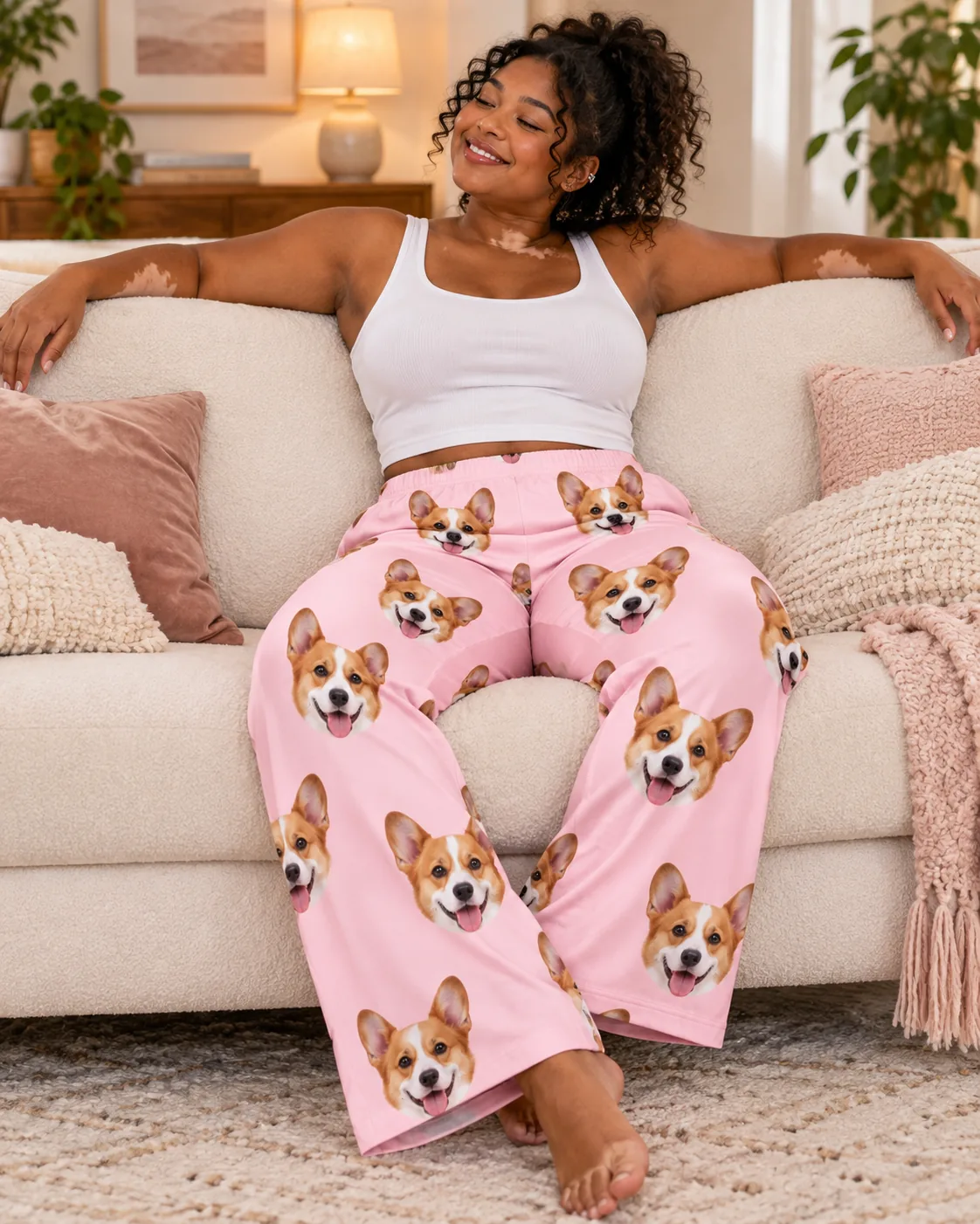 Woman stretching out on a cream sofa in blush pink pajama pants printed with smiling corgi faces