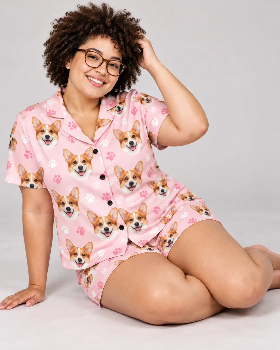 Smiling woman in glasses sitting on a light gray studio floor in a blush pink paws corgi pj set