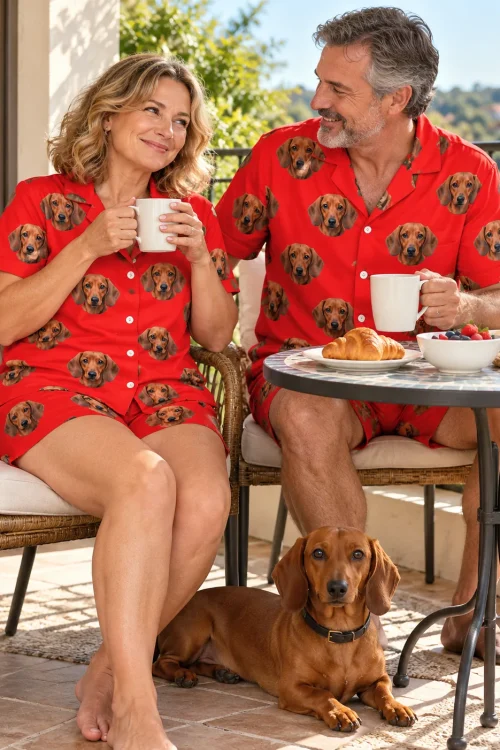 Couple sharing breakfast on a sunny patio in red dachshund pajama sets, their dachshund lying below