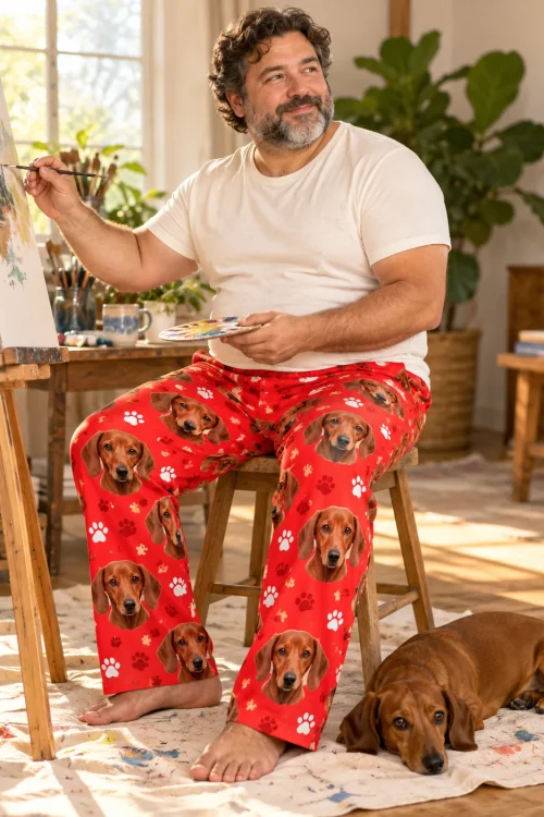 Bearded man painting at an easel while his dachshund rests on the drop cloth, in red paws dachshund pajamas