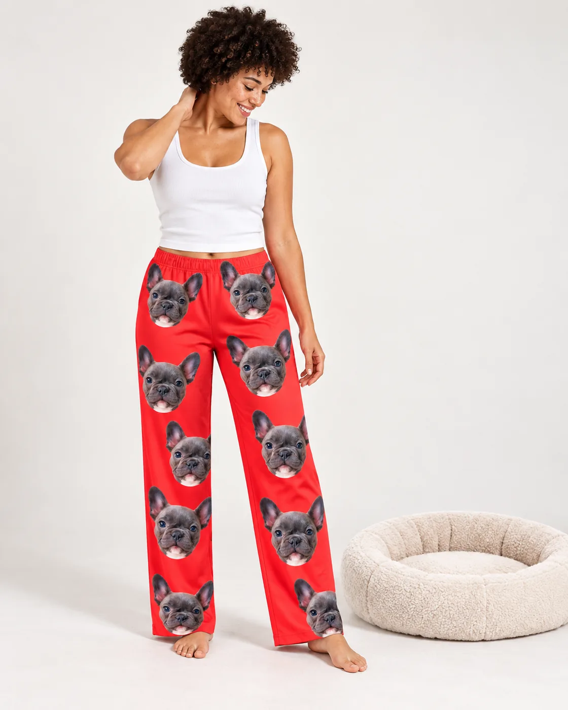 Pet face pajamas: woman with curly hair beside a dog bed in red pajama pants printed with French bulldog faces