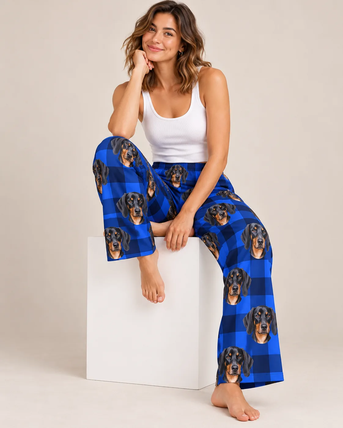 Woman sitting on a white cube in blue plaid doberman pajama pants, hand under her chin, studio