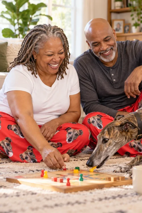 Couple playing a board game on a rug in red greyhound pajamas while their brindle greyhound sniffs the pieces