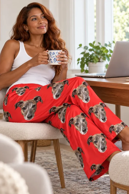 Woman with a blue-and-white mug at a sunny desk, feet up in red greyhound pajamas printed with dog faces