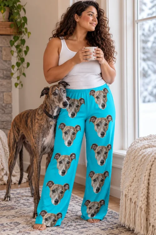 Woman with a mug by a snowy window in teal greyhound pajamas, her greyhound leaning on her near a fireplace