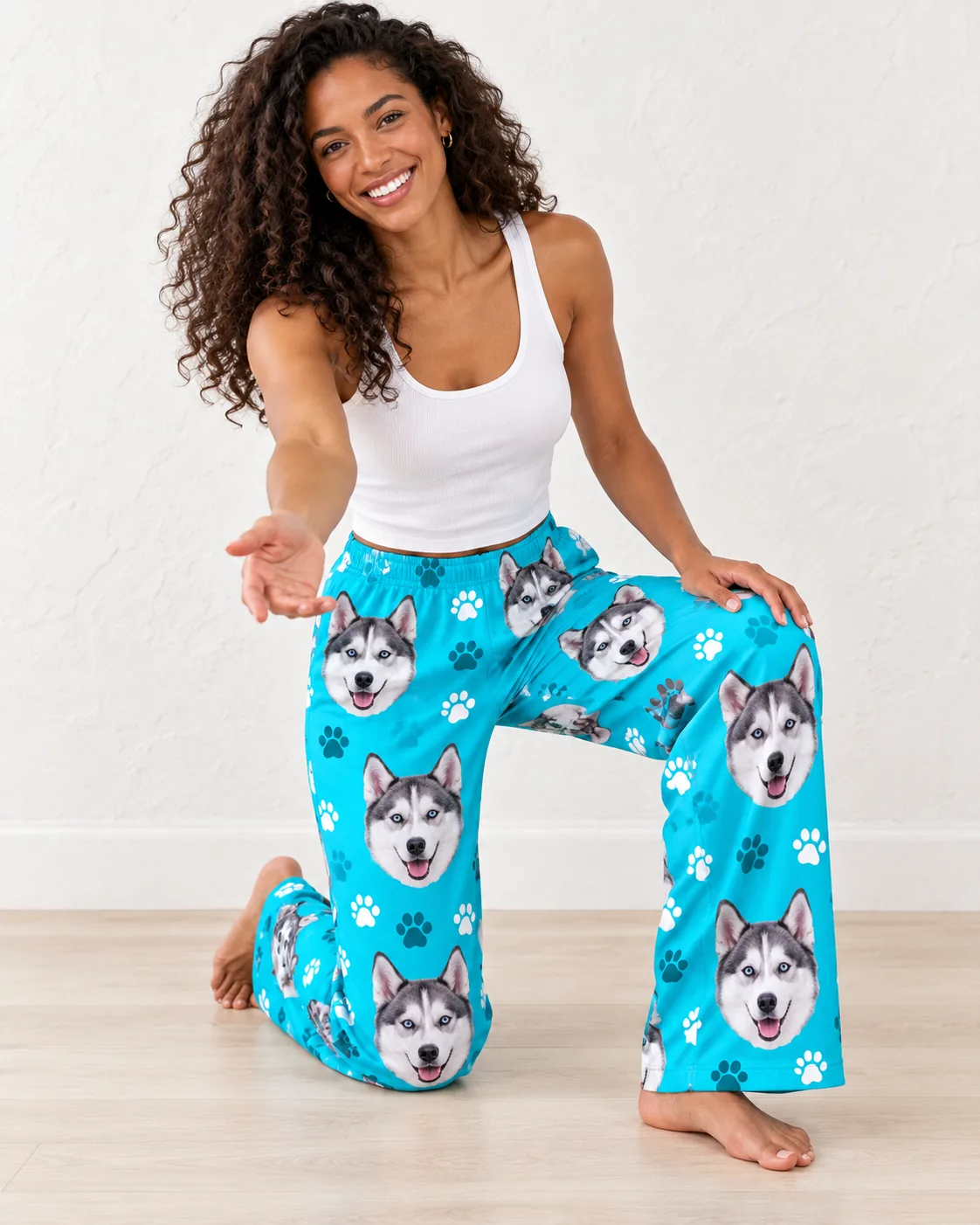 Woman kneeling on a light floor in teal husky dog pajama pants with paw prints and husky faces, hand outstretched