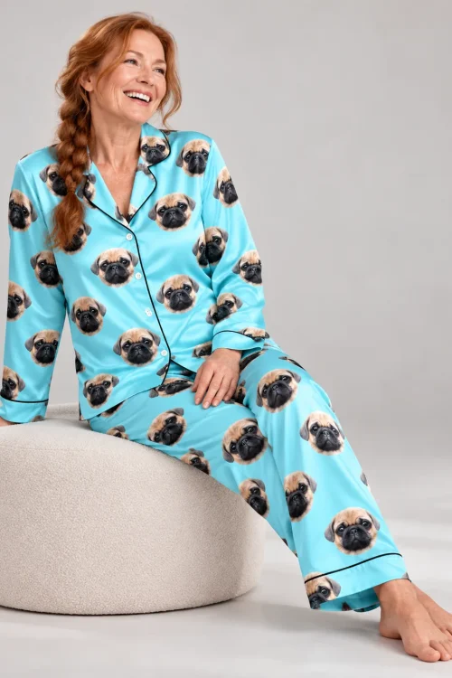 Smiling woman with a long red braid seated on a pouf in light blue satin pajamas with pug faces