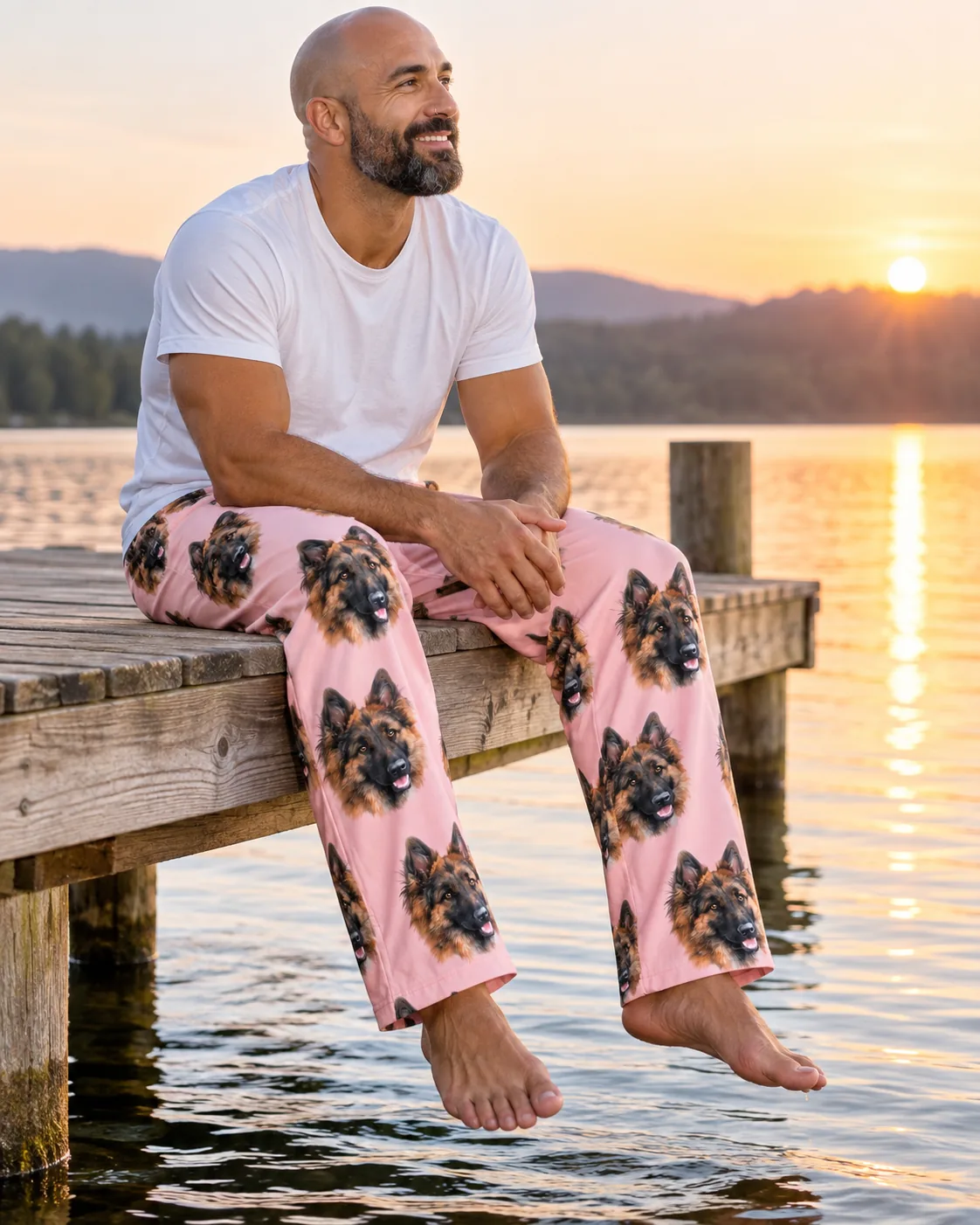 Man sitting on a lake dock at sunrise in light pink pajama pants printed with German Shepherd faces