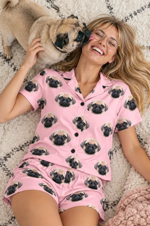 Woman in glasses lying on a rug in a light pink pajama set while her pug licks her cheek
