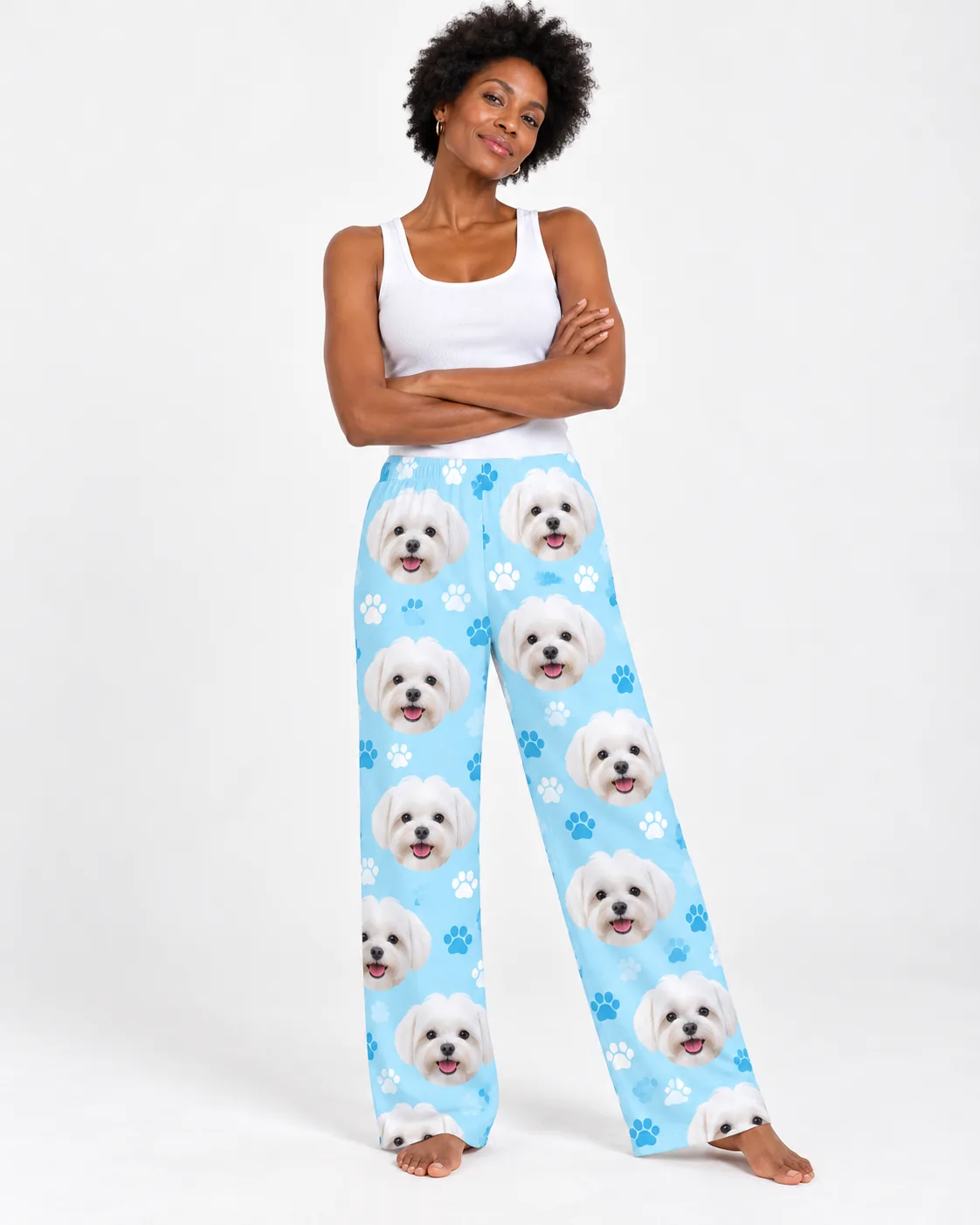 Smiling woman with arms crossed in a white tank and light blue paws maltese pjs, barefoot against a white studio wall