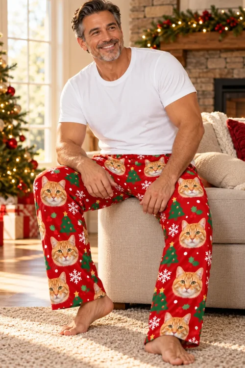 Smiling man on a sofa arm by the Christmas tree in red cat pajama pants with snowflakes