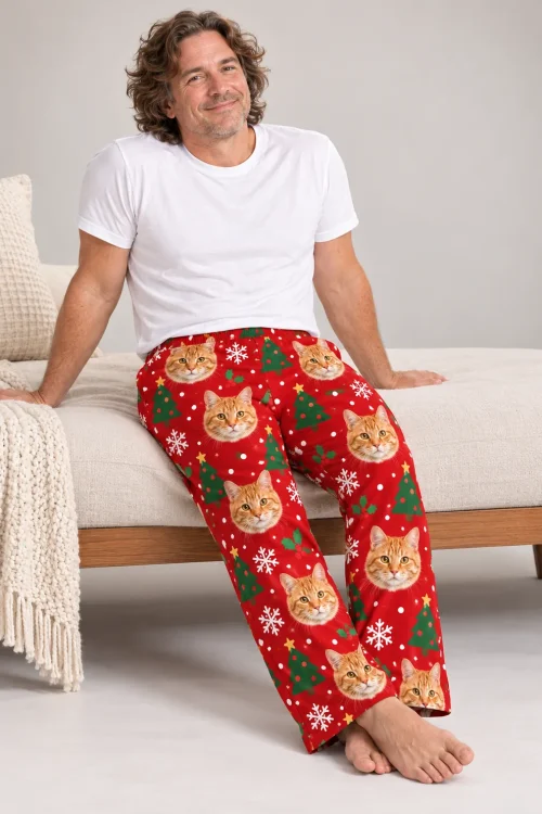 Curly-haired man on the edge of a daybed in red Christmas cat pajamas with trees and snowflakes