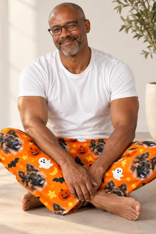 Man sitting cross-legged in orange mens Halloween pajama pants with German Shepherd faces and pumpkins