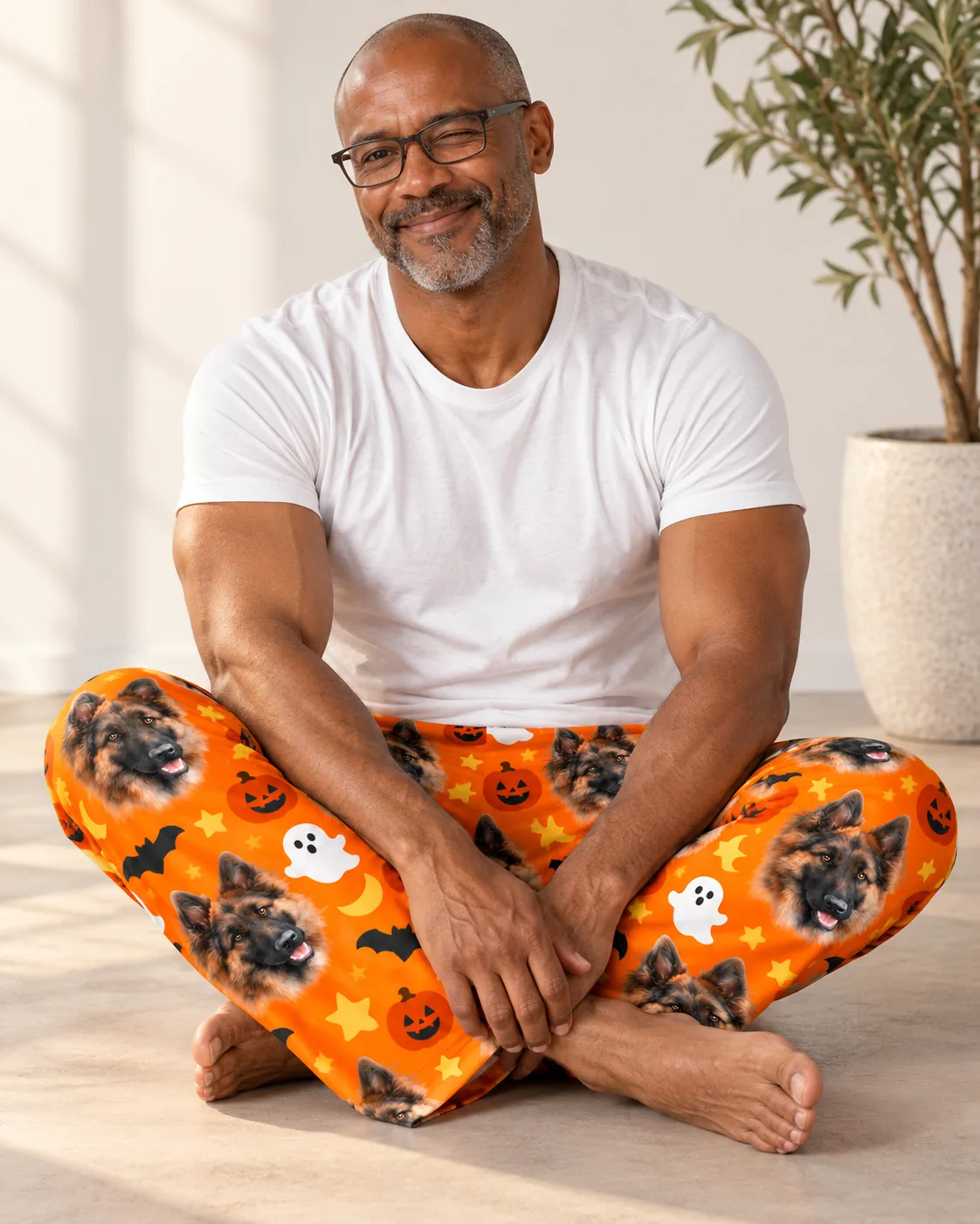 Man sitting cross-legged in orange mens Halloween pajama pants with German Shepherd faces and pumpkins
