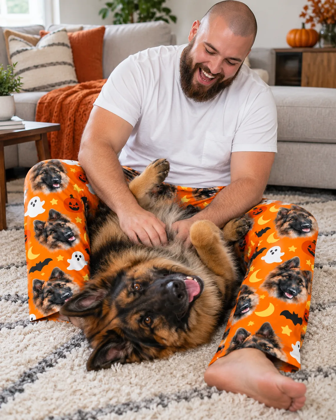 Bearded man rubbing his German Shepherd's belly while wearing orange Halloween pajama pants with its face