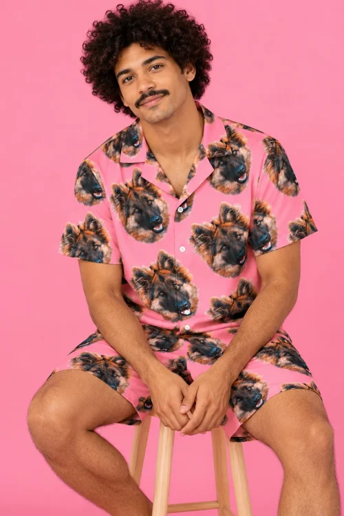 Man on a wooden stool against pink, wearing a pink shirt and shorts with German shepherd faces