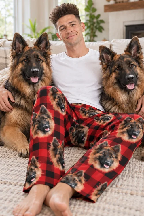 Man on the floor between two German Shepherds, wearing red plaid pajama pants with their faces