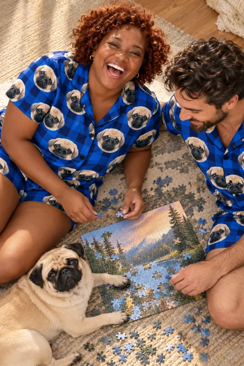 Laughing couple doing a jigsaw puzzle on a rug in blue plaid pug face pajama sets beside their fawn pug