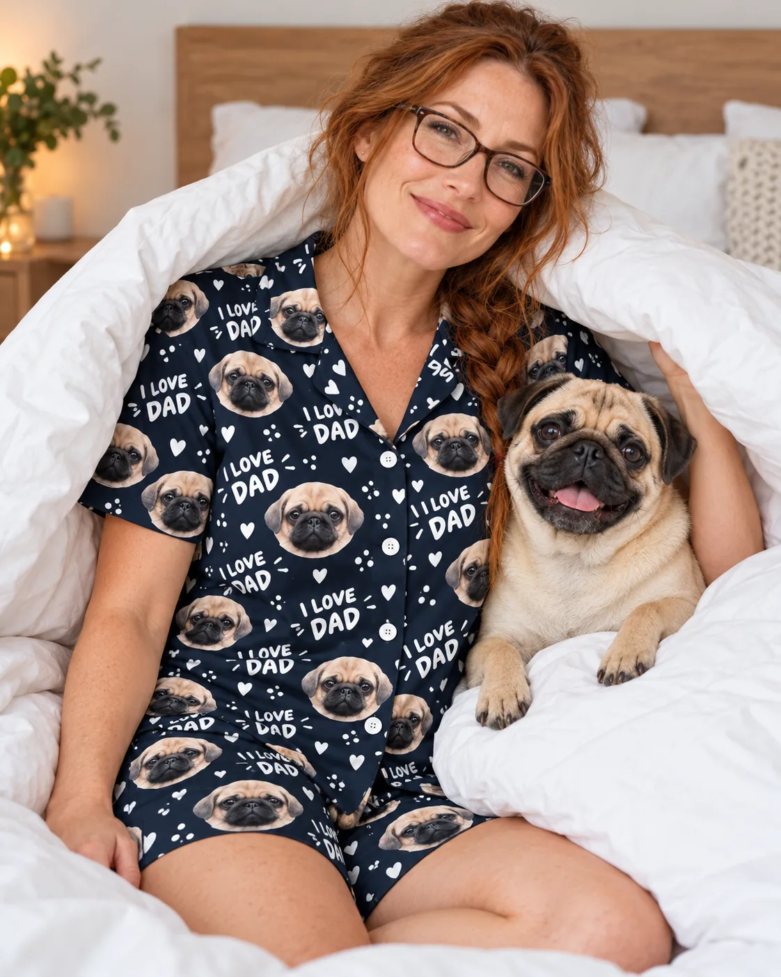 Woman in glasses snuggles under a white duvet with her fawn pug, wearing a navy I Love Dad pug face pajama set