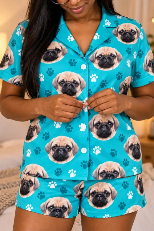Close-up of woman buttoning a teal paw print short pajama set printed with fawn pug faces in a lamp-lit bedroom