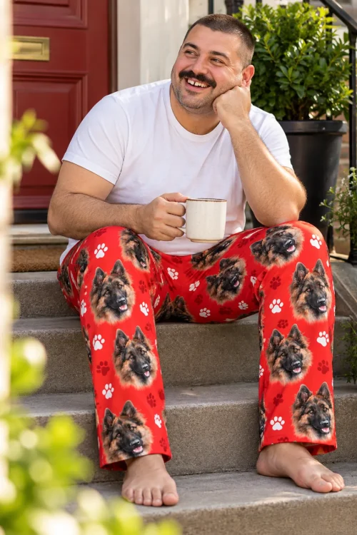 Man with a mug sits on front steps in red paws pajama pants printed with German shepherd faces