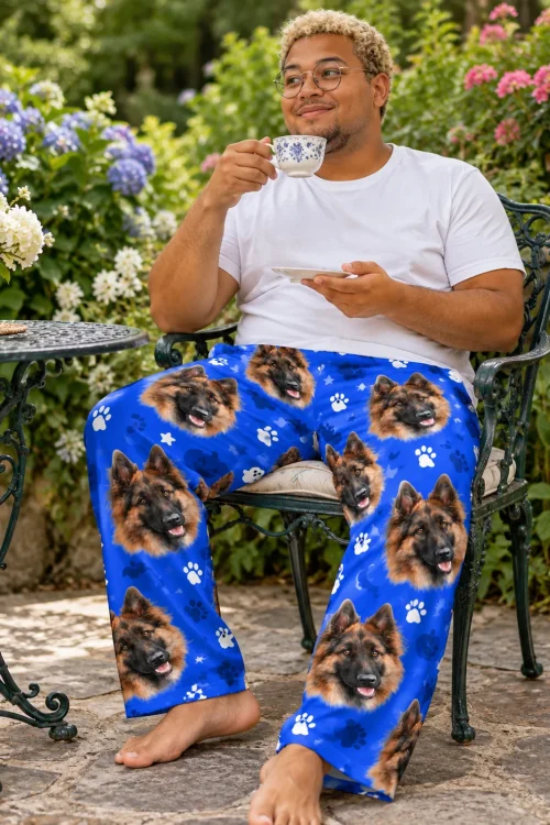 Man sips tea in a flower garden in royal blue paws pajama pants printed with German shepherd faces