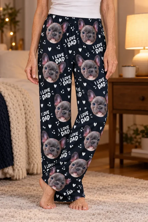Close-up of navy I Love Dad pajama pants with grey French bulldog faces and white hearts in a warm lamp-lit bedroom