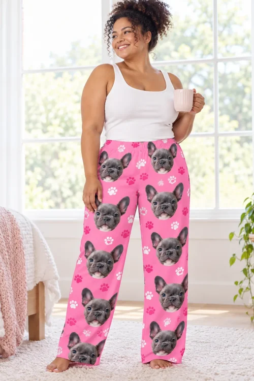 Smiling woman with a pink mug by a bright bedroom window in pink paw print pajama pants with French bulldog faces