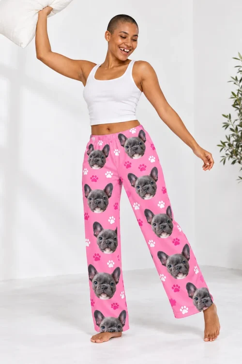 Joyful woman swinging a pillow in pink paw print pajama pants with French bulldog faces, white studio with a plant