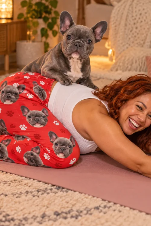 Laughing red-haired woman on a yoga mat in red paw print pajama pants as her grey French bulldog climbs on her back