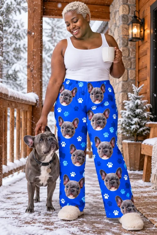 Woman with a mug pets her grey French bulldog on a snowy cabin porch, wearing royal blue paw print pajama pants