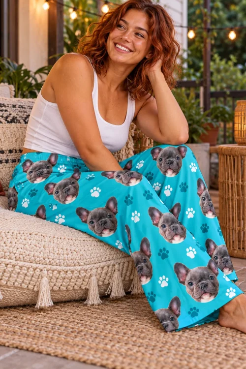 Smiling red-haired woman on a patio floor cushion under string lights in teal paw print French bulldog pajama pants