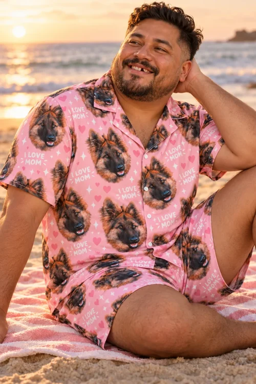 Smiling man sitting on a beach towel at sunset in a pink I Love Mom short pajama set printed with German shepherd faces