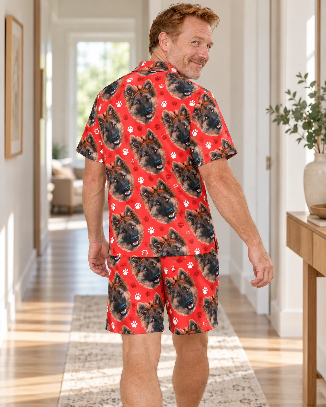 Back view of smiling man in a sunny hallway in a red paw print short pajama set printed with German shepherd faces