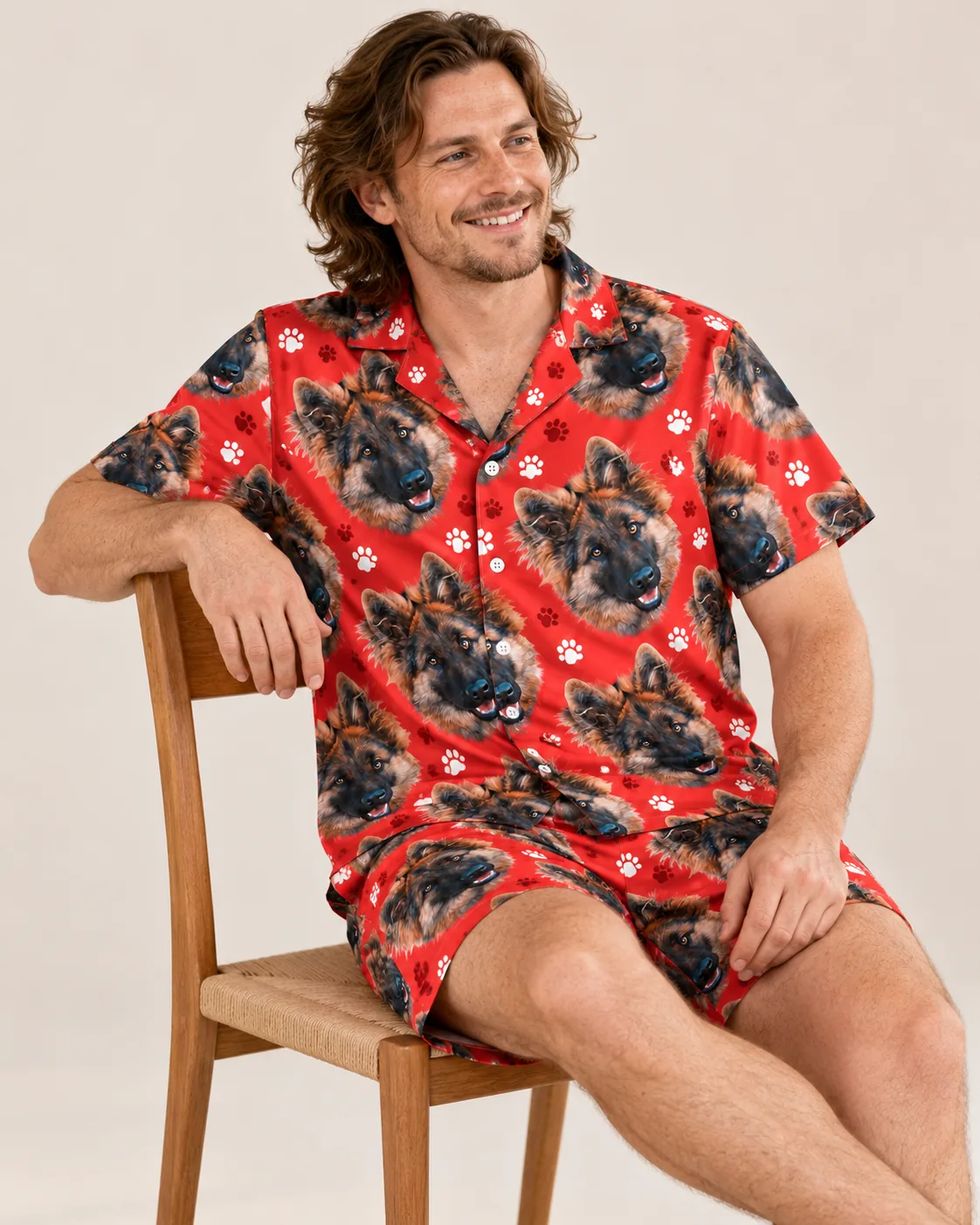 Smiling man on a wooden chair in a red paw print short pajama set with German shepherd faces, light studio backdrop